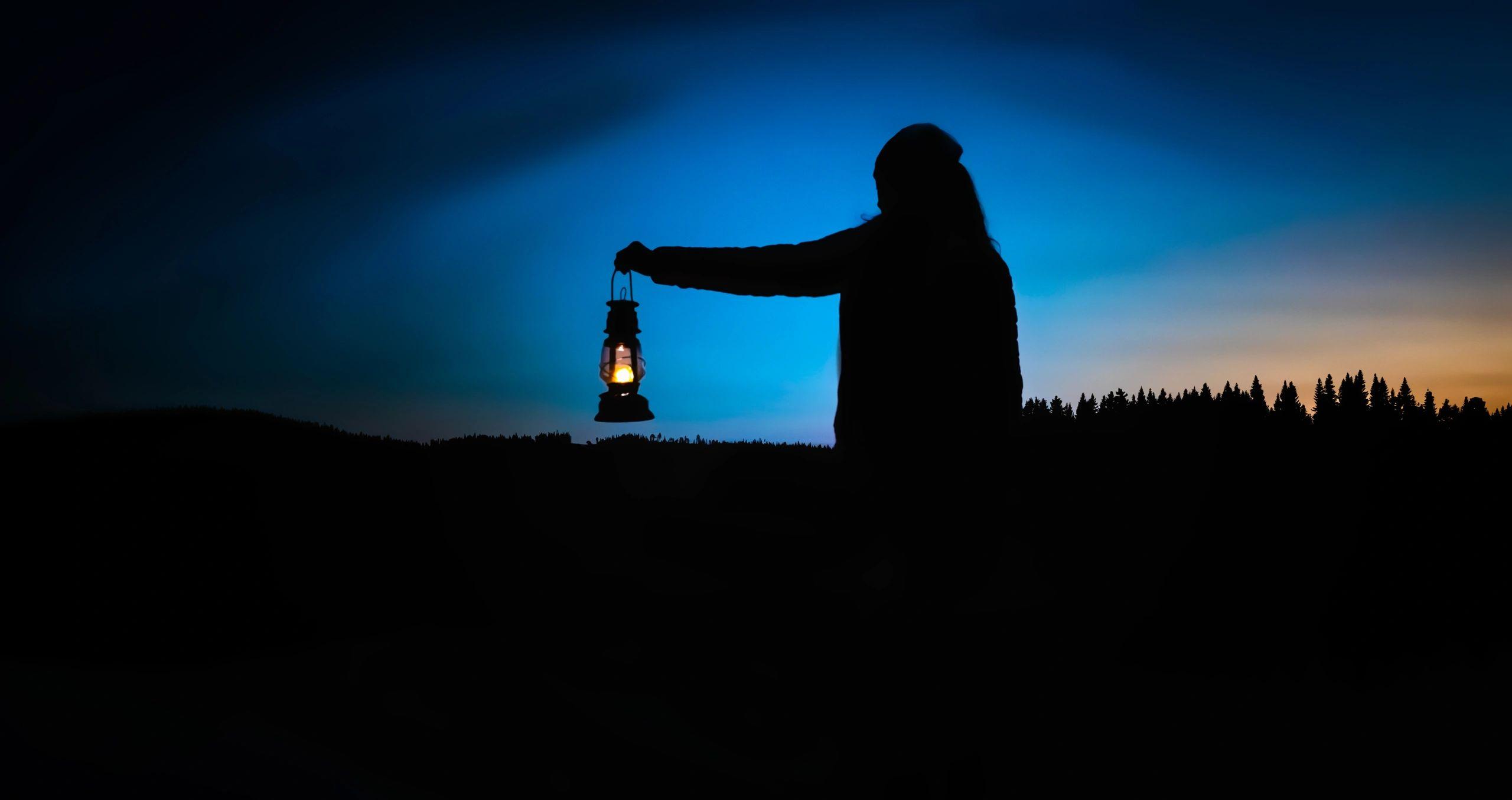 A silhouette of a person holding a glowing lantern against a deep blue dusk sky