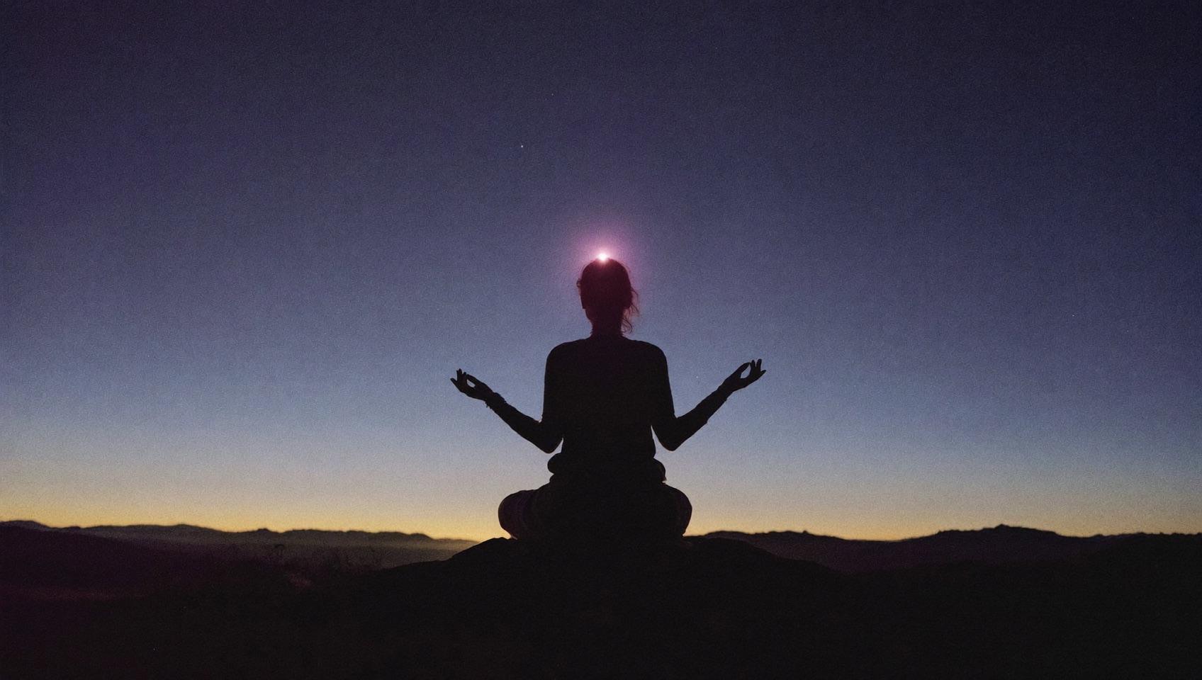 A woman seated in meditation on a hilltop at twilight with soft light above her head